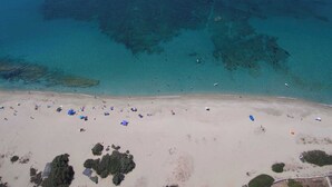 Ein Luftbild zeigt einen Strand mit klarem türkisblauem Wasser, Sandufer und vereinzelten Sonnenschirmen.