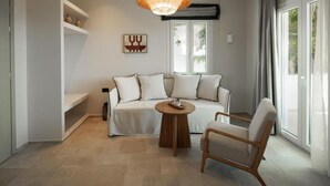 Ein modernes Wohnzimmer mit einer Couch, einem runden Couchtisch und einem Sessel. Ein Fenster mit Vorhängen, ein Wandregal mit Dekoration und eine Deckenleuchte sind ebenfalls zu sehen.