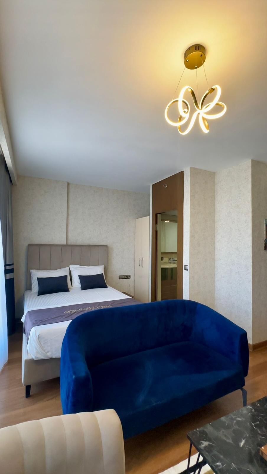 Una habitación de hotel con una cama, un sillón azul, una mesita pequeña y una lámpara de techo moderna.