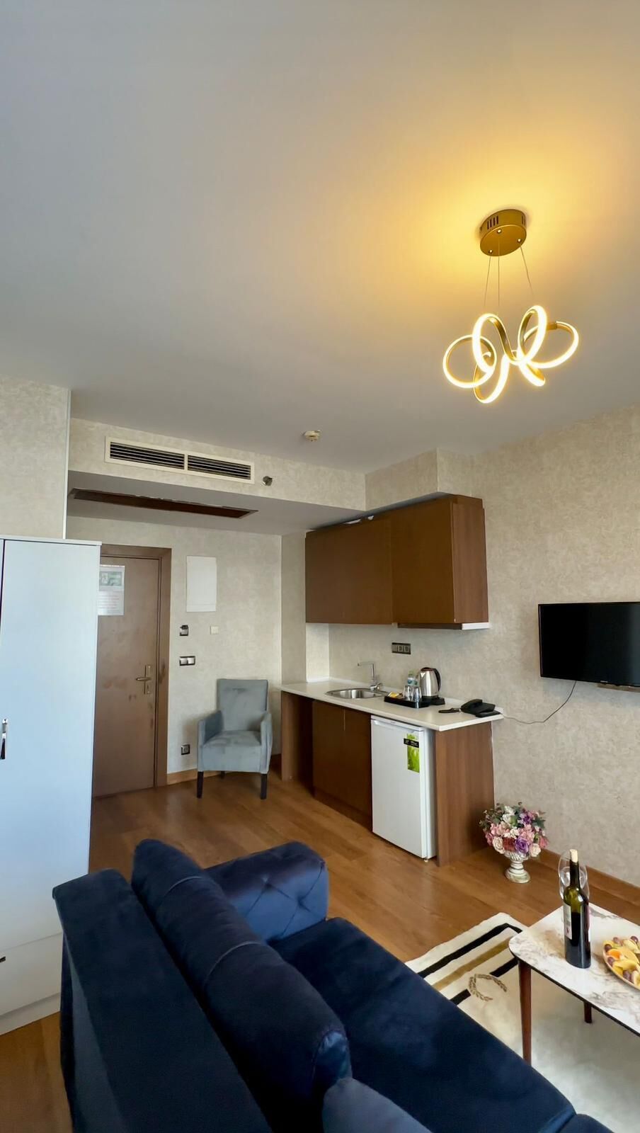 Een compacte woonruimte met een kitchenette, een bank, een klein tafeltje met een fles en een aan de muur bevestigde televisie.