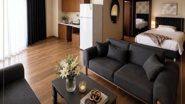 A modern hotel room with a sofa, a coffee table with flowers, a bed, a kitchen area, and a mirror.