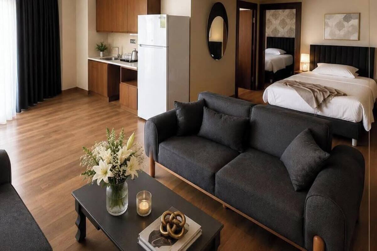A modern hotel room with a sofa, a coffee table with flowers, a bed, a kitchen area, and a mirror.