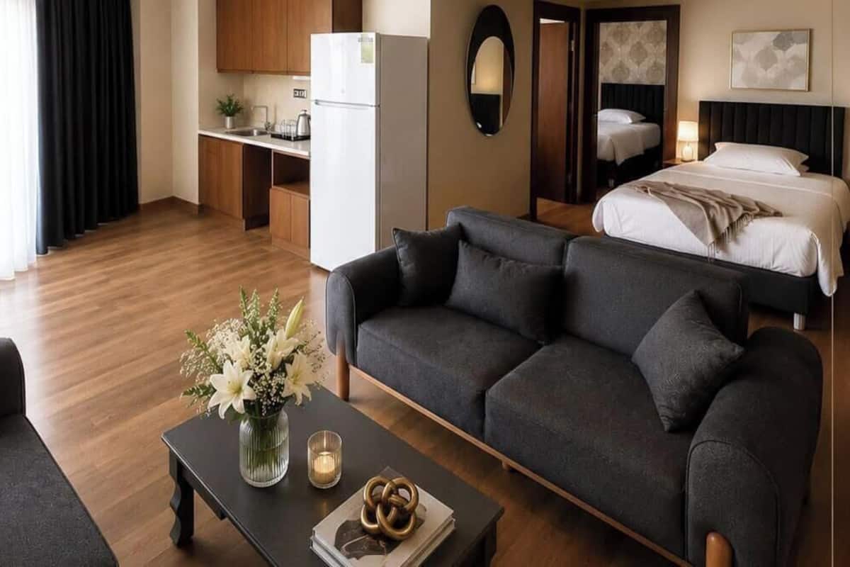 A modern hotel room with a sofa, a coffee table with flowers, a bed, a kitchen area, and a mirror.