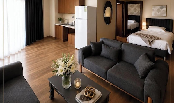 A modern hotel room with a sofa, a coffee table with flowers, a bed, a kitchen area, and a mirror.