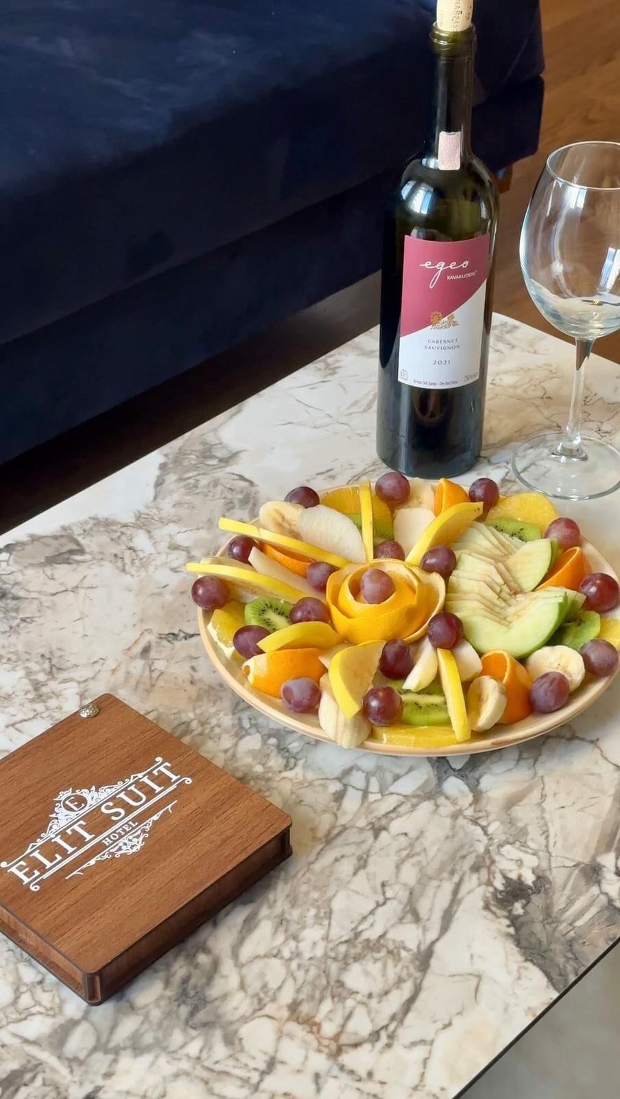 A bottle of wine, a glass, and a fruit platter on a marble table.