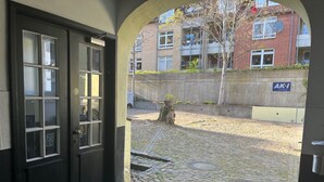 Een met stenen bestraat binnenhof met een zwarte deur, een betonnen muur en een gebouw met het bord 'AKI Immobilien' op de achtergrond.