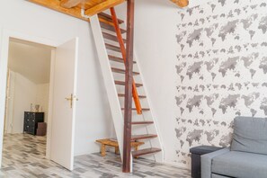 Une pièce avec un escalier en bois, un canapé gris, un papier peint représentant une carte du monde et un sol carrelé.