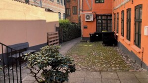 Un estrecho callejón entre edificios, con un pequeño jardín, una banca y un contenedor de basura.