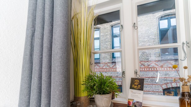 A window with a view of a brick building, a potted plant, a tray with candles, and a sign on the windowsill. - fewo1846 - Matrosenkoje - Centrally Located Apartment in an Older Building for 2 People on the 2nd Floor (Flensburg)