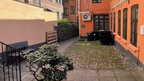 A narrow alleyway between buildings with a small garden area, a bench, and a trash bin.