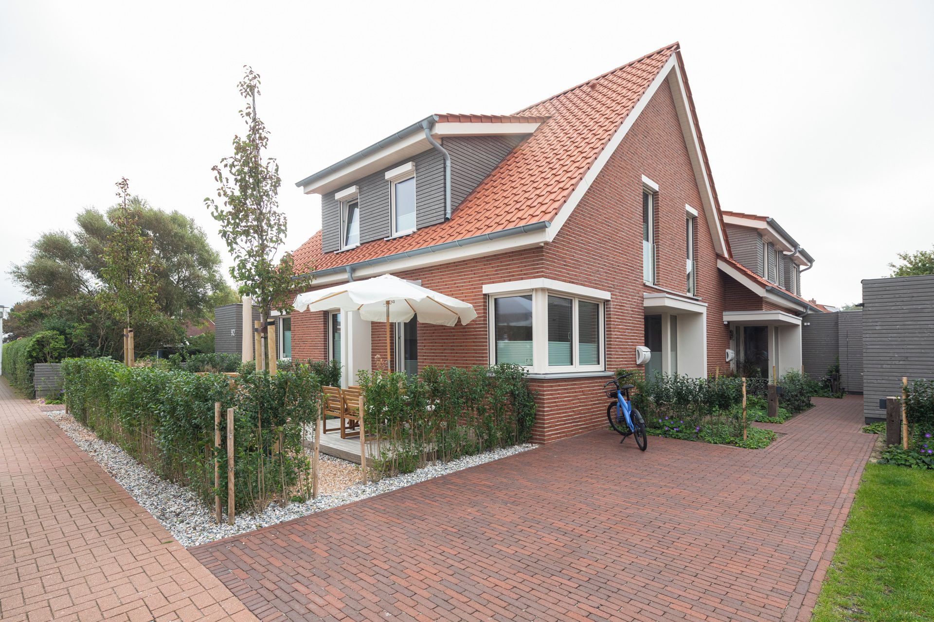 Une maison à deux étages avec un toit recouvert de tuiles rouges, une terrasse couverte avec une table et des chaises, et un vélo garé à l’extérieur.