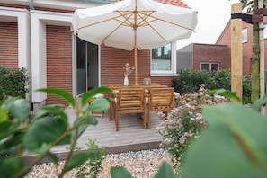 A patio with a wooden table and chairs, a white umbrella, and a brick house in the background. - // DUNE NEIGHBORHOOD NO. 1 // (Norderney)