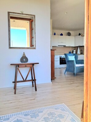 A room with a wooden floor, a table with a lamp, a chair, a kitchen area with cabinets, and a mirror on the wall. - A stone's throw from the sea! (Cannigione)