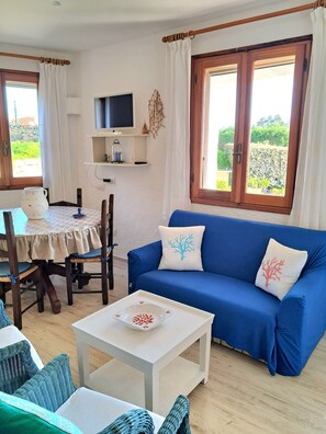 Una sala de estar con un sofá azul, una mesa de centro blanca, sillas de mimbre, una mesa de comedor y un televisor sobre una estantería.