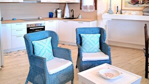 Een moderne keuken met witte kastjes, een fornuis en een spoelbak. Er staan twee blauwe rieten stoelen met gestreepte kussens bij een witte tafel met een schaal.