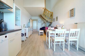 A modern kitchen with white cabinetry, a wooden staircase, and a dining area with white chairs. - Residence Ostseestrand Apartment 29 (Scharbeutz)
