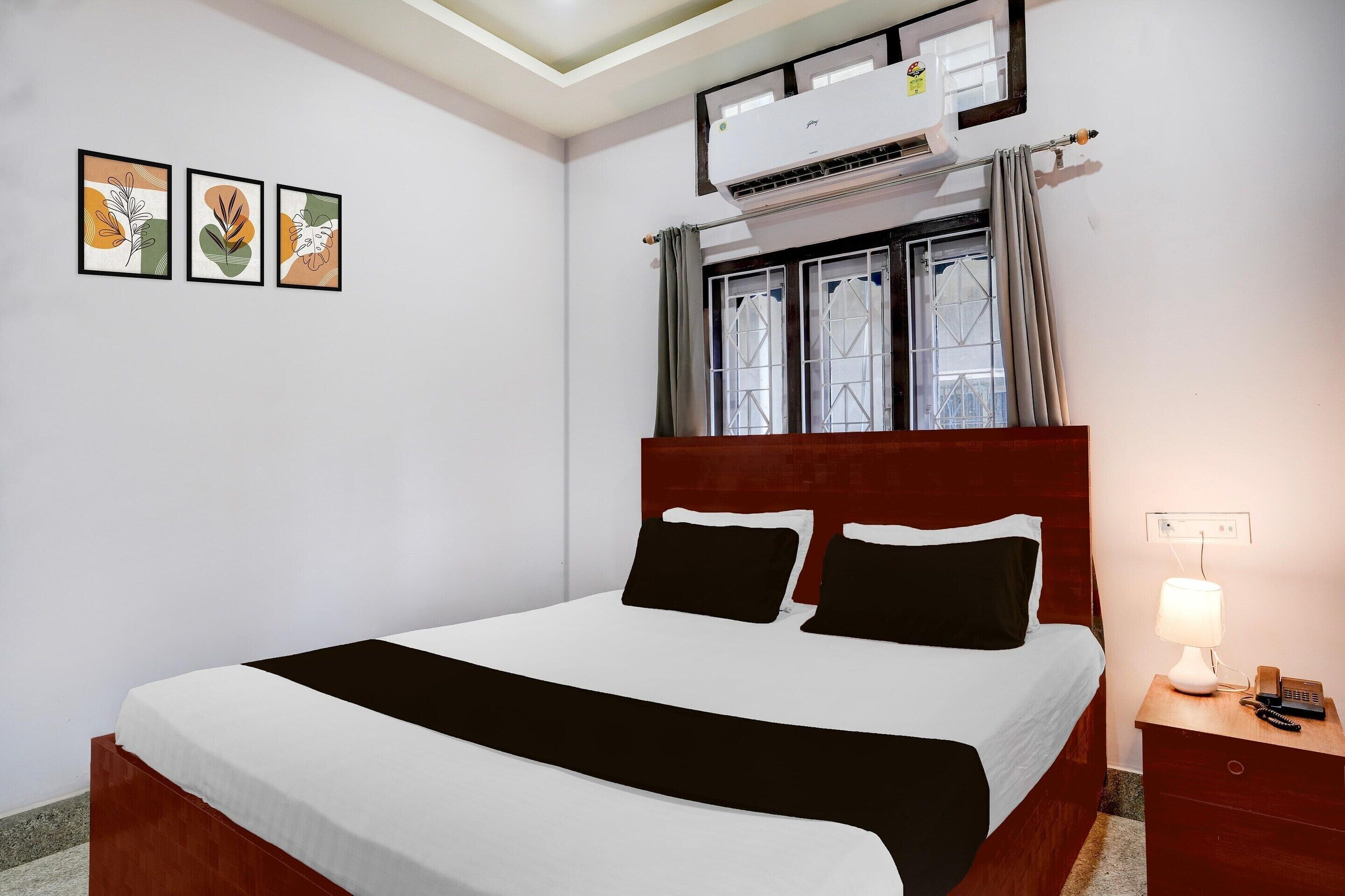 Ein Hotelzimmer mit einem Bett, einem Nachttisch mit Lampe, einer Klimaanlage und drei gerahmten Kunstwerken an der Wand.