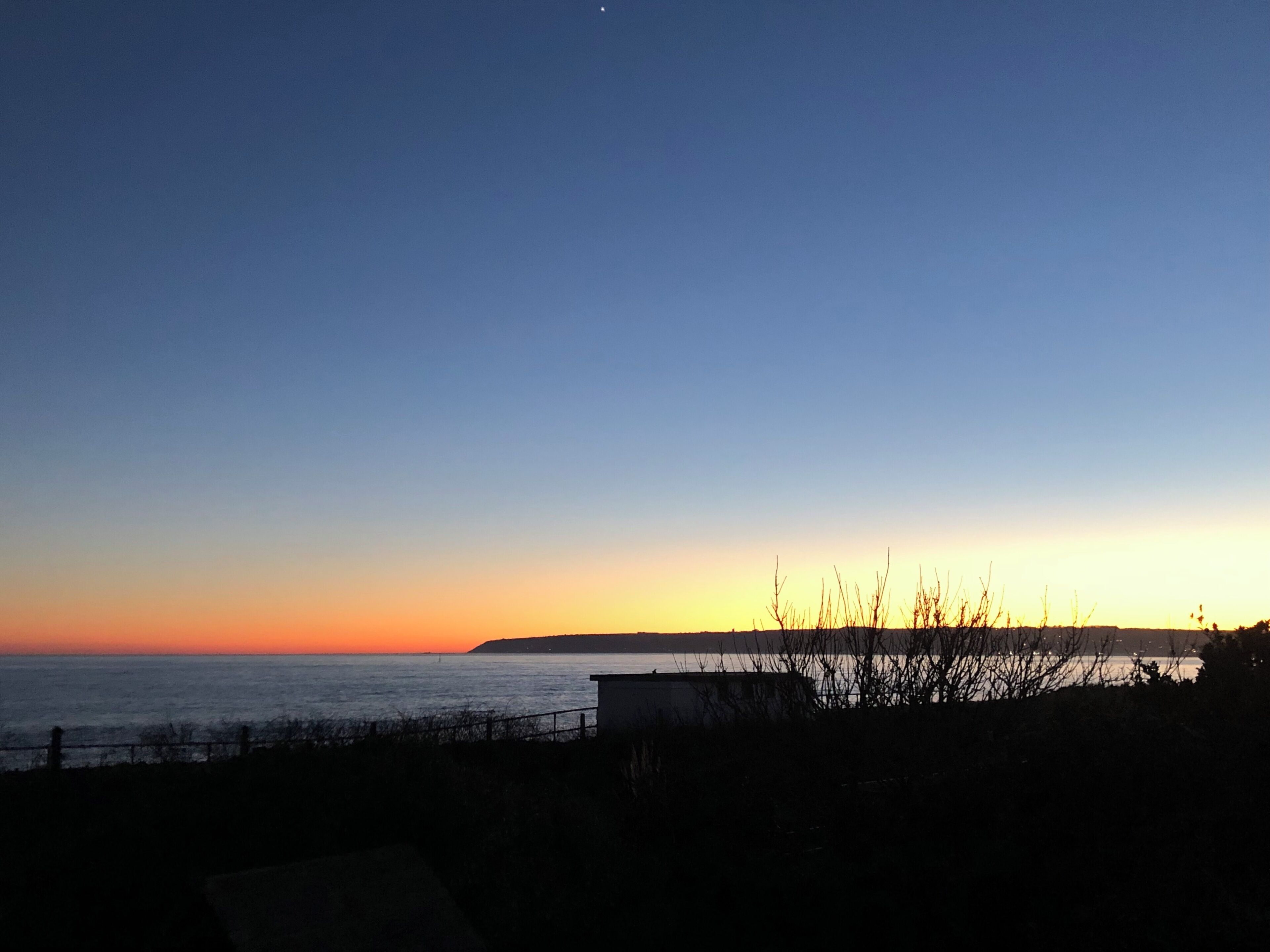 A coastal sunset view with a silhouette of a building and vegetation in the foreground.