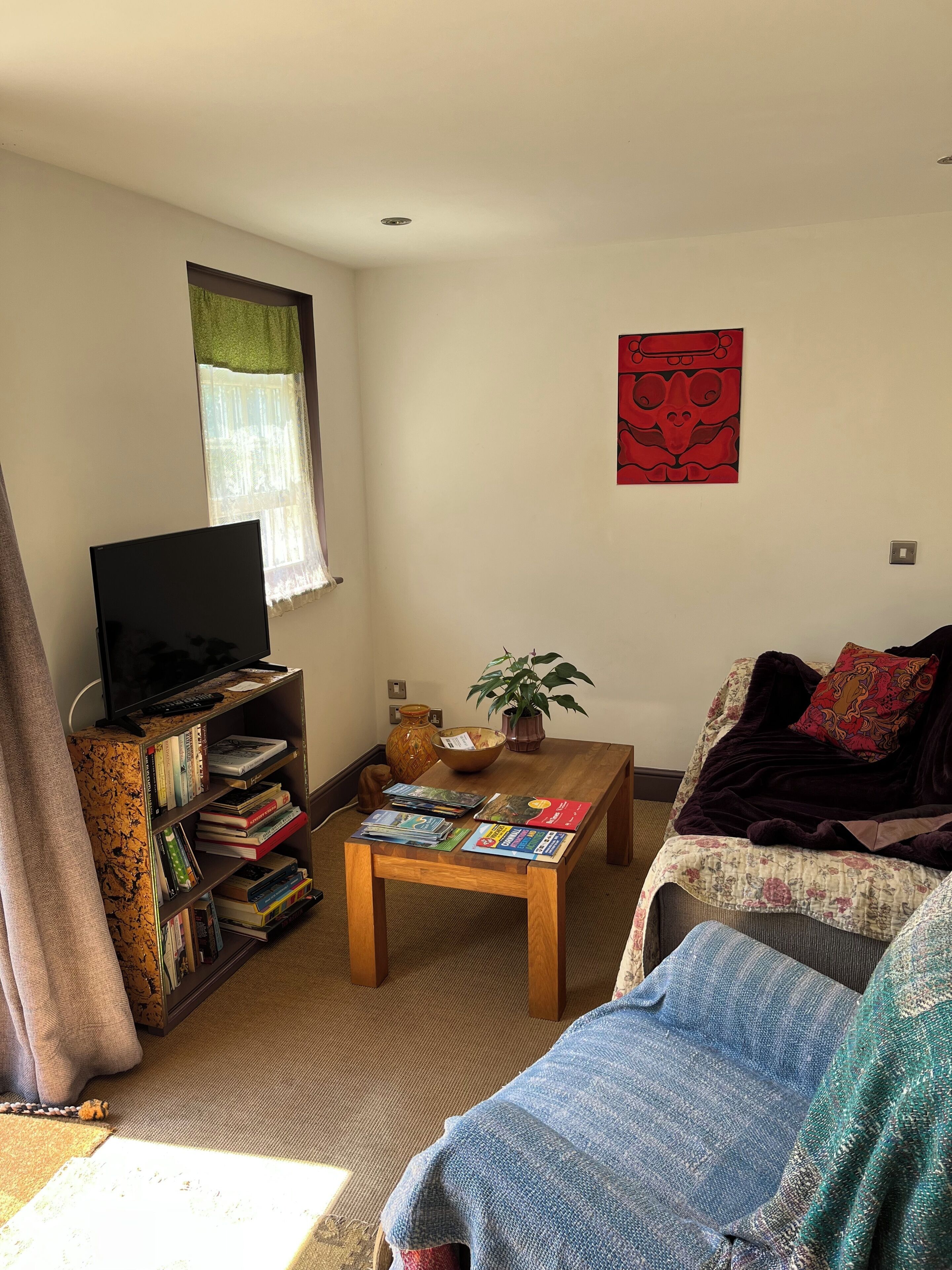 A living room with a TV on a shelf, a coffee table with magazines, a couch with a blanket, and a wall with a red artwork.
