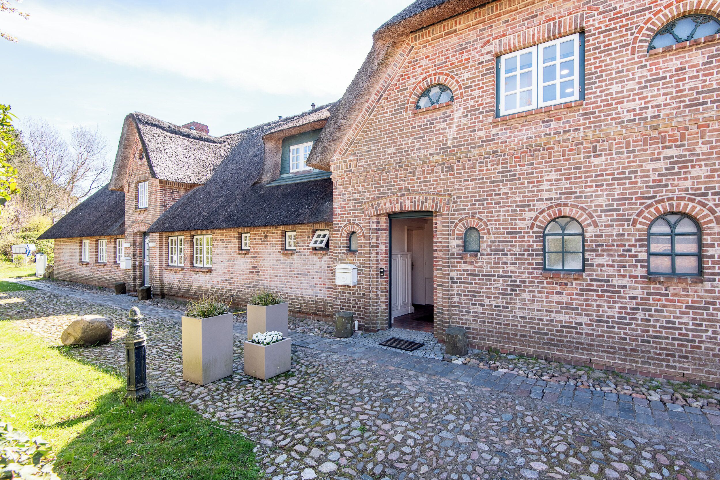 A brick building with a thatched roof, a stone-paved courtyard, and a large planter with flowers.
