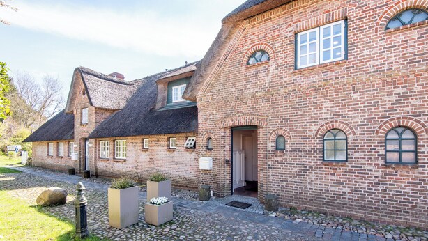 Ein Backsteinhaus mit einem Strohdach, einem gepflasterten Innenhof und einem großen Blumenkübel.