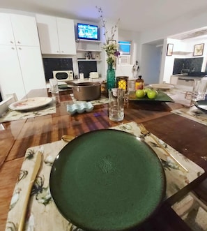 Mesa de jantar com pratos, talheres, uma tigela, um vaso com flores e uma televisão ao fundo.
