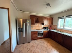 A kitchen with wooden cabinets, a stainless steel refrigerator, a built-in oven, and a window with a view of greenery. - Spacious Provencal villa with swimming pool and pétanque court (Saint Paul En Foret)