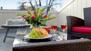 Ein Außenbereich mit Sitzgelegenheit, einem Tisch, Korbstühlen, einer Glasplatte, einer Vase mit Blumen, einem Teller mit Obst und zwei Gläsern mit Getränken.