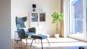 Ein modernes Wohnzimmer mit einem grauen Polsterstuhl, einem kleinen Beistelltisch, einer Topfpflanze, einem Schrank mit Glastüren und einem großen Fenster.