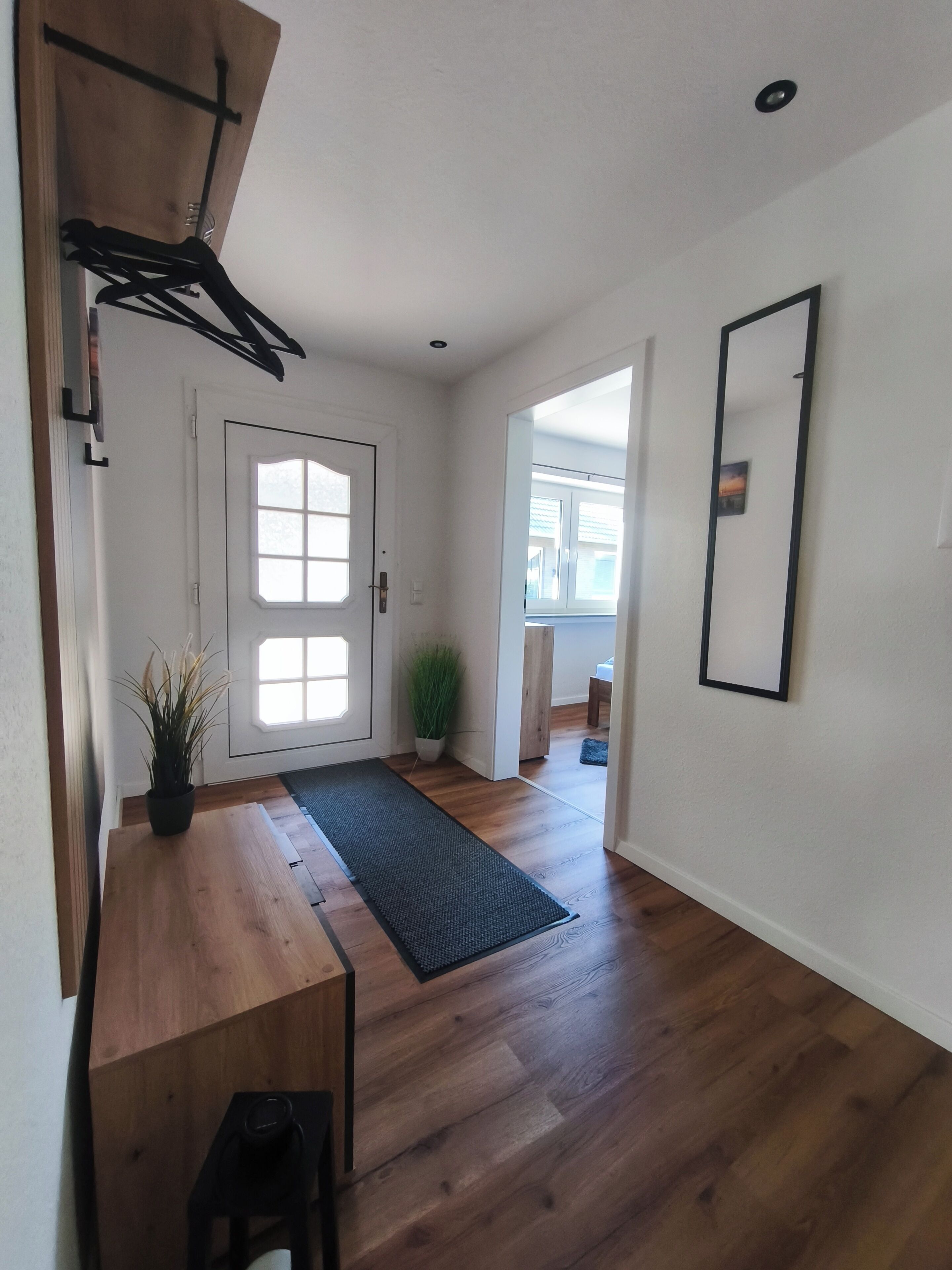 Un couloir avec un plancher en bois, une porte dotée d’une fenêtre, une plante en pot, un banc et un miroir accroché au mur.
