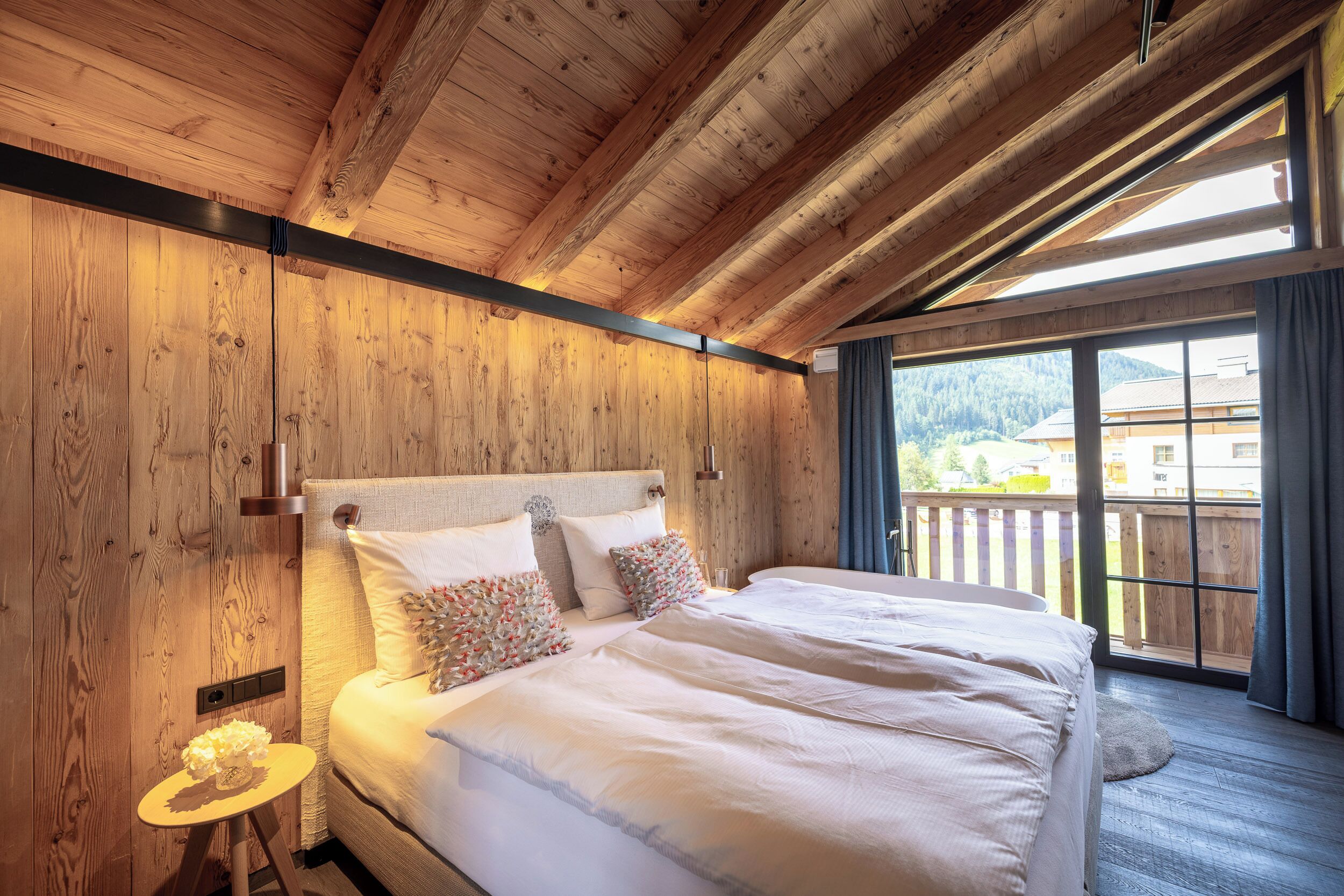 Ein Schlafzimmer mit Holzvertäfelung, einem großen Bett, einem Nachttisch und einem Fenster mit Blick auf Gebäude und Bäume.