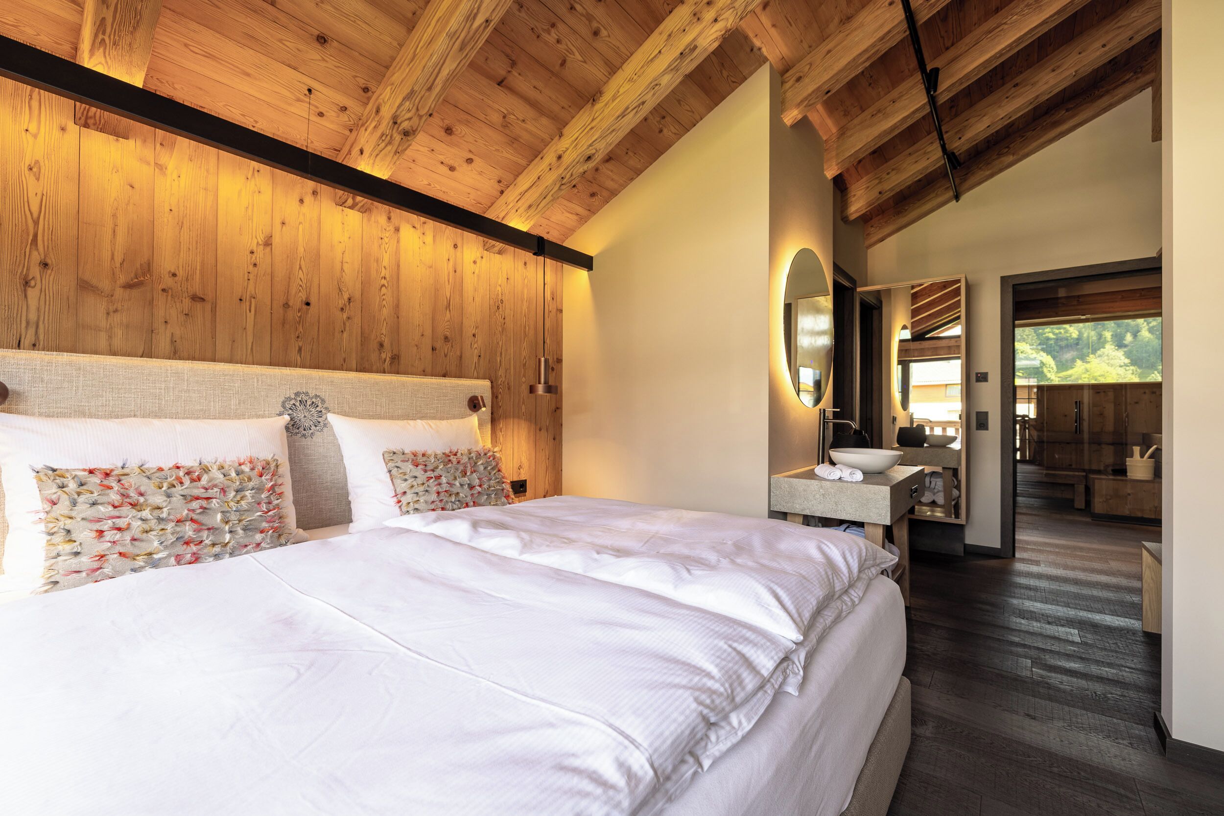 Ein Schlafzimmer mit Holzboden, einem großen Bett mit weißen Leinentüchern, einem Nachttisch mit Spiegel und Blick auf einen Wald durch ein Fenster.