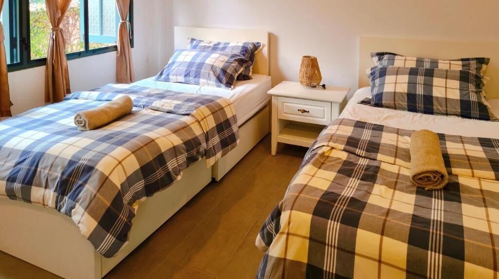 A room with two beds, each with a checkered bedspread and a rolled towel. There is a nightstand with a lamp between the beds.