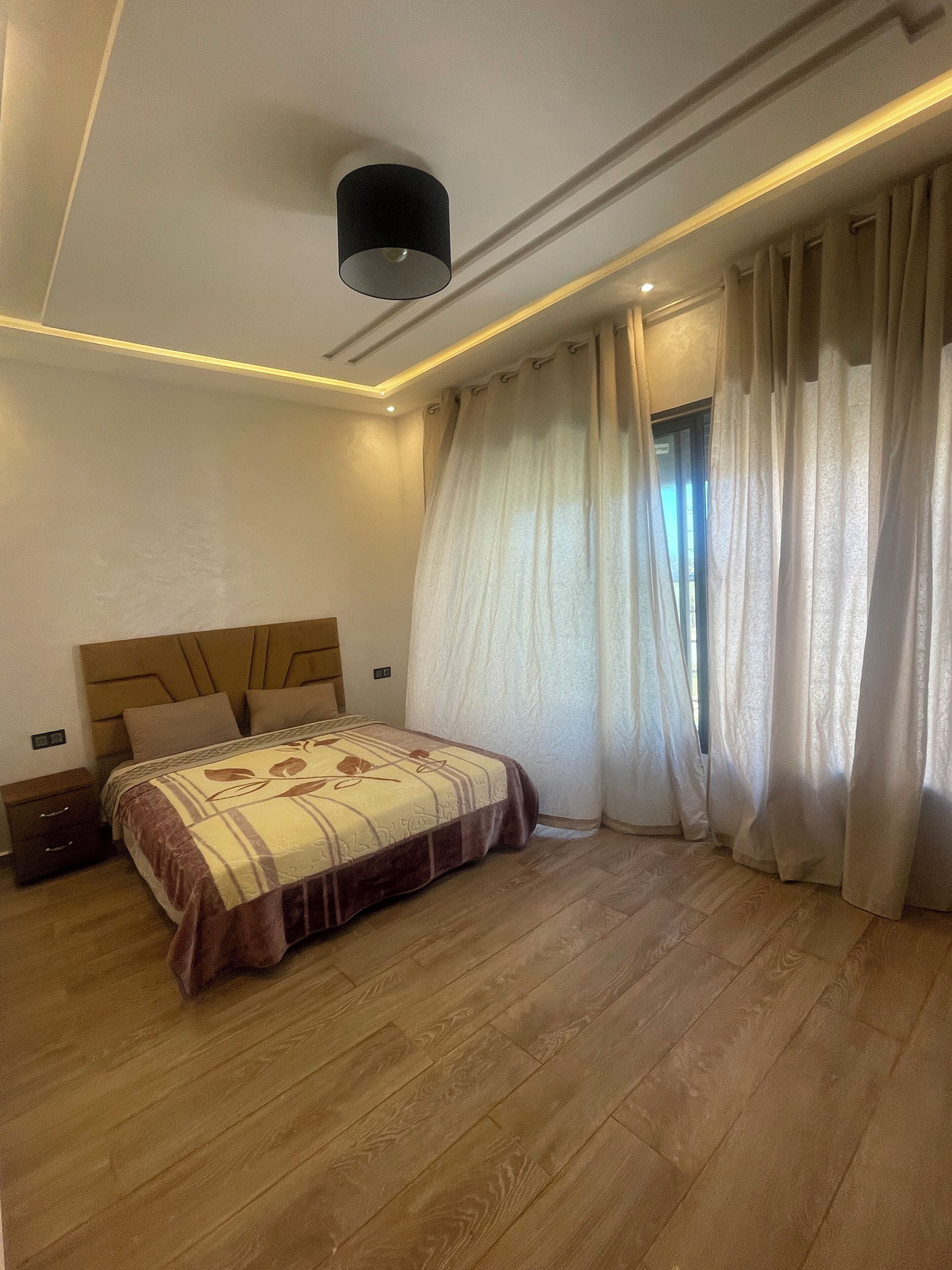 Un dormitorio con una cama, una mesita de noche, una lámpara y una ventana con cortinas.