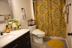 Une salle de bain équipée de toilettes, d’un lavabo, d’un miroir, d’un porte-serviettes et d’un rideau de douche à motifs floraux jaunes.