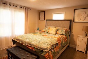 Une chambre à coucher avec un lit, une table de chevet, une lampe, une fenêtre avec des rideaux et un tableau encadré au mur.