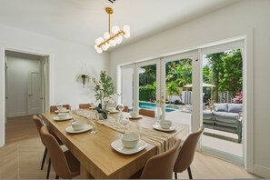A dining area with a wooden table set for four, surrounded by chairs, with a view of a swimming pool and outdoor seating. - Fox Eden Resort; 8bd, Pool& Spa Oasis (Fort Lauderdale)