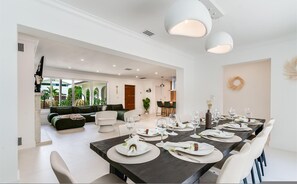 A modern dining area with a dark wooden table set for a meal, white chairs, and a view of a living room with a green sofa and a dining area with a chandelier. - Fox Eden Resort; 8bd, Pool& Spa Oasis (Fort Lauderdale)