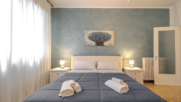 Een keurig opgemaakt bed met een blauwe spreideken en twee opgerolde handdoeken, een schilderij aan de muur, twee nachtkastjes met lampen, en een deur met geëtst glas.