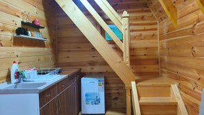 A wooden cabin interior with a kitchen area, a small refrigerator, and a wooden staircase.