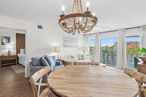 Een ruime woonkamer met een ronde houten tafel, een witte bank met blauwe kussens, een kroonluchter en een groot raam met uitzicht op de omgeving.