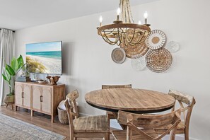 A dining area with a round wooden table, wooden chairs, a chandelier, and a wall-mounted TV. - New at Maravilla • Gulf View Balcony • Free Beach Service • Sleeps 8 (Miramar Beach)