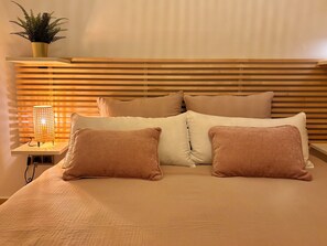 Una cama con cabecera de madera, una mesita de noche con lámpara y una planta en maceta.