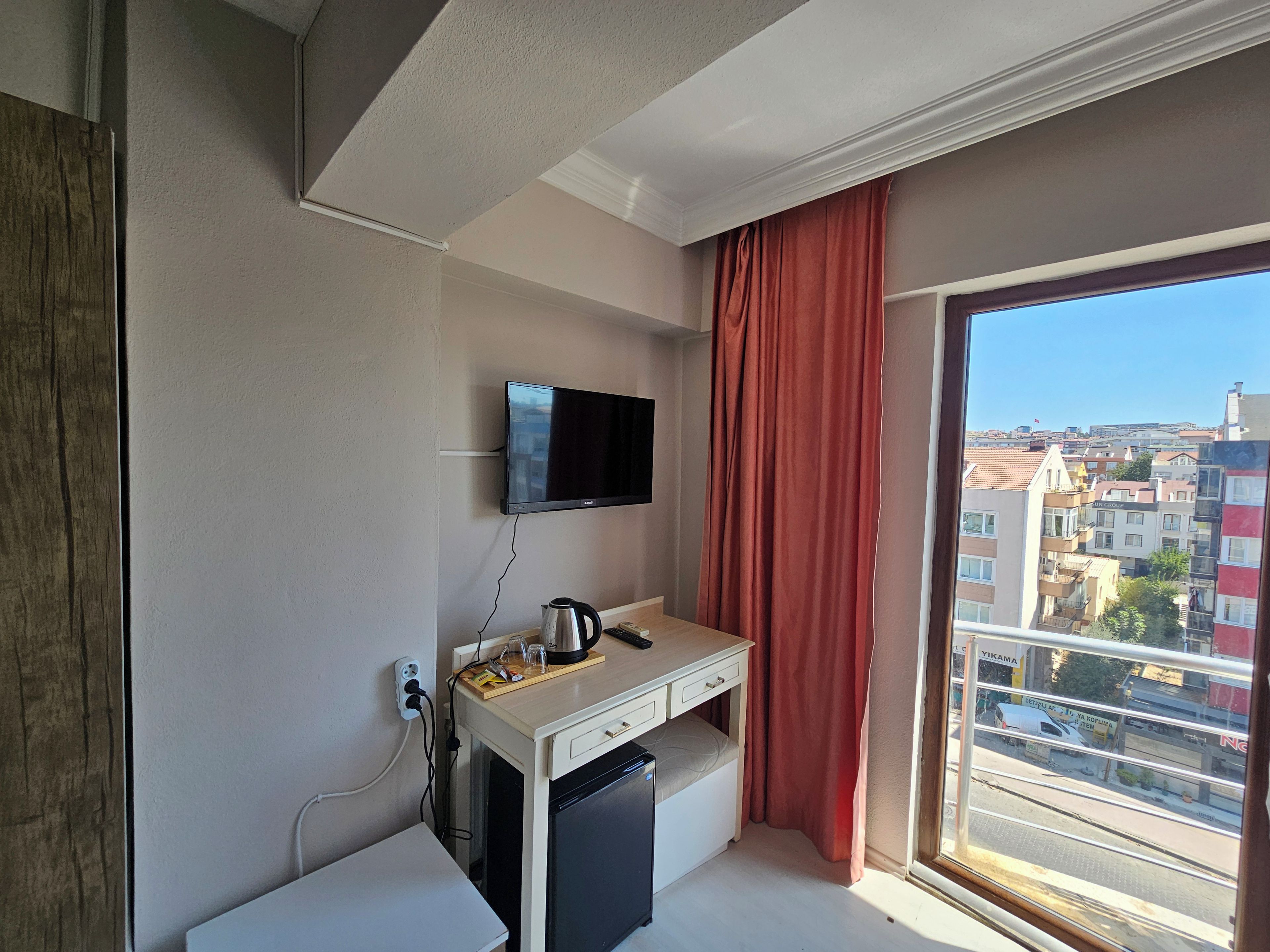 A compact room with a flat-screen TV mounted on the wall, a small kitchenette with a coffee maker, and a window with a view of the cityscape.