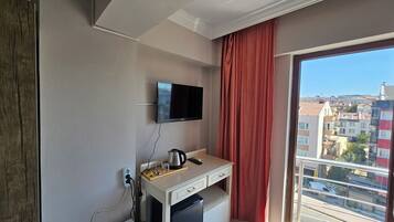 A compact room with a flat-screen TV mounted on the wall, a small kitchenette with a coffee maker, and a window with a view of the cityscape.