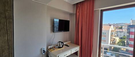 A compact room with a flat-screen TV mounted on the wall, a small kitchenette with a coffee maker, and a window with a view of the cityscape.