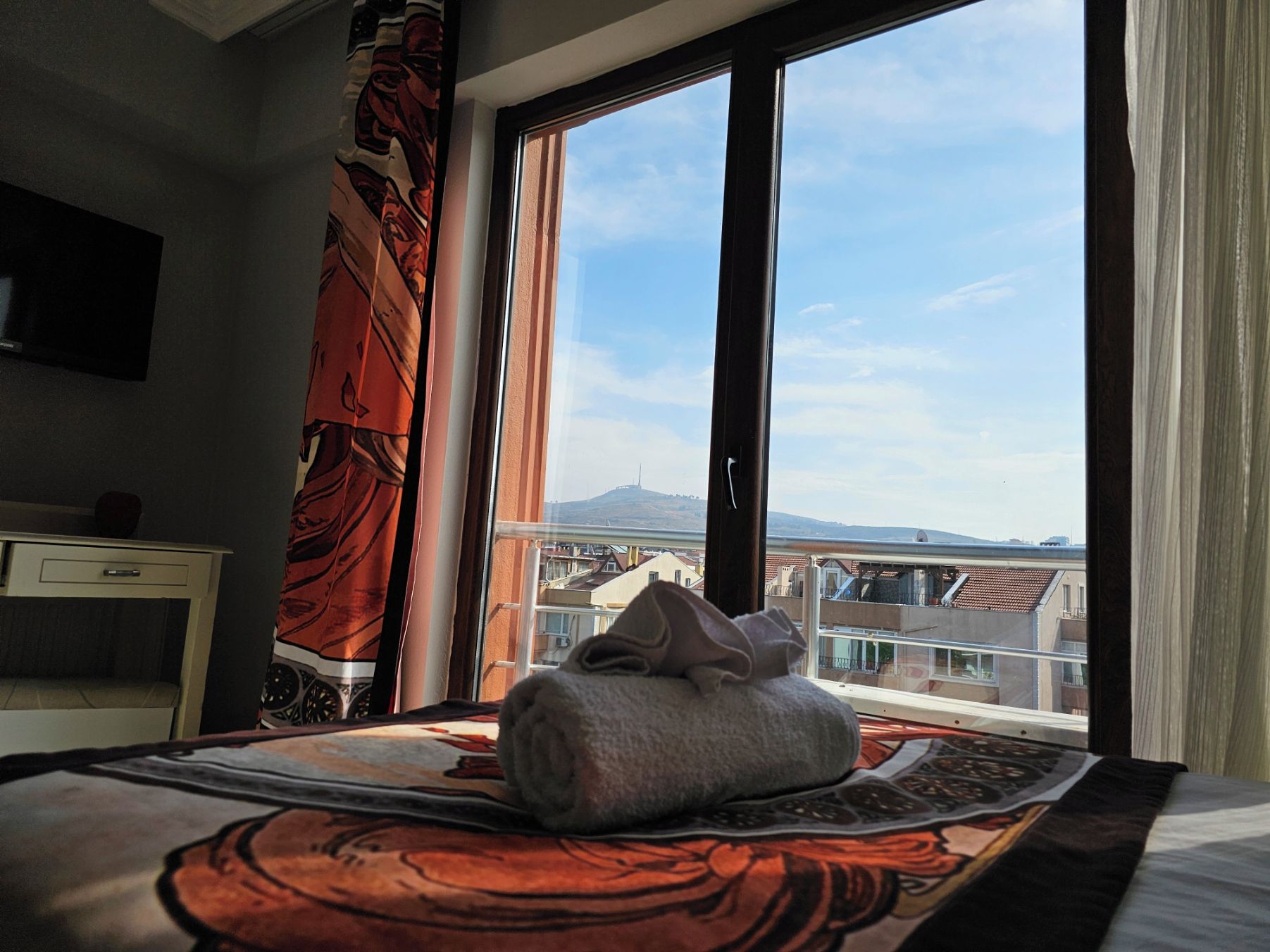 Ett hotellrum med en säng, ett skrivbord och ett stort fönster med utsikt över byggnader och en kulle.