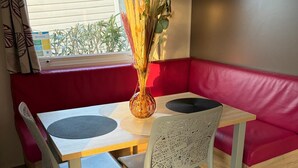 Uma área de refeições com mesa, cadeiras, um vaso com plantas secas, um banco estofado vermelho e um aparelho de ar condicionado.
