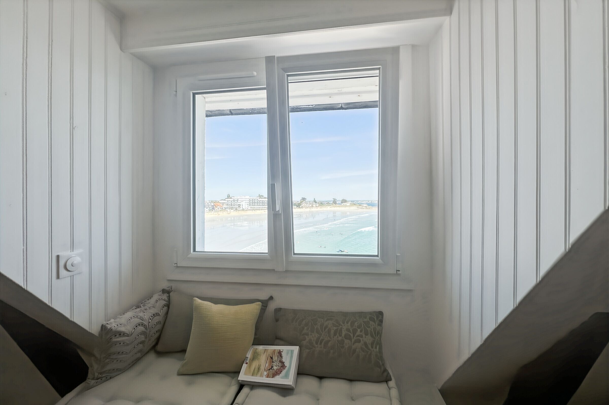 A room with a window overlooking a beach, a cushioned bench with pillows, and a book on the bench.