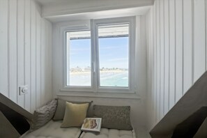 Una habitación con ventana que da a una playa, un banco acolchado con almohadones y un libro sobre el banco.
